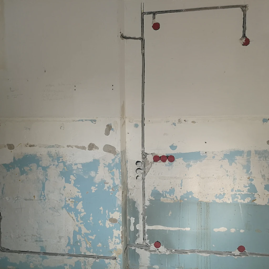 Eine teilweise gestrichene Wand mit freiliegenden elektrischen Leitungen und roten Steckdosen; die Wand weist Abnutzungserscheinungen und Flecken mit alter blauer und weißer Farbe auf.